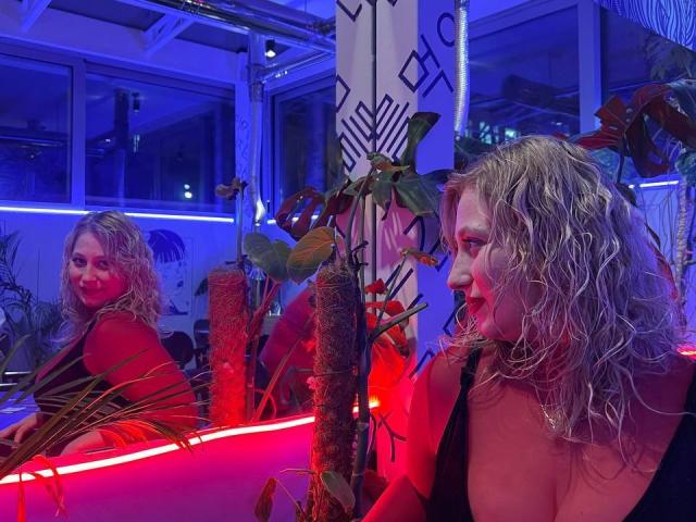 SerenadeAlice with wet blonde hair, red top, and glowing red lipstick, sits in a blue-lit club, reflected in a mirror, with red and blue lights on a plant.