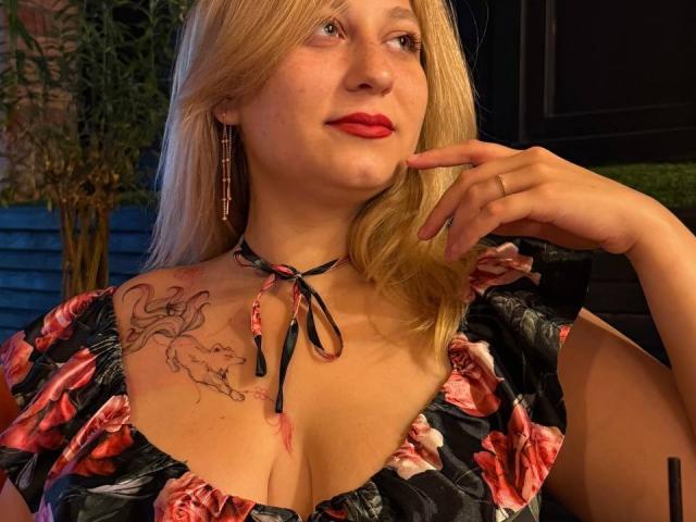 SerenadeAlice has blonde hair, red lipstick, and a floral dress with a low neckline, showing her tits and a tattoo on her left shoulder. She's pointing to her chin.