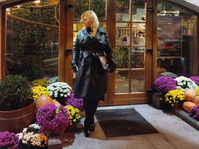 SerenadeAlice with blonde hair, black leather coat, black boots, holding purse, standing in front of wooden glass door, surrounded by colorful potted flowers.