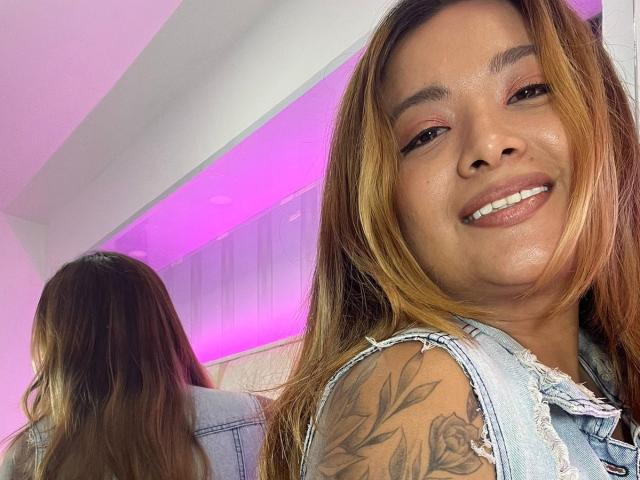 BellaLumi, smiling with her nose slightly upturned, has light brown skin, long blonde and brown hair, and a rose tattoo on her right shoulder. Her denim vest is visible.