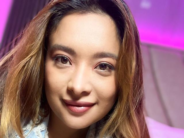 BellaLumi's face, showing brown eyes, subtle makeup, and a slight smile. She has light brown skin, shoulder-length blonde hair with darker roots, and is wearing a white lace top.