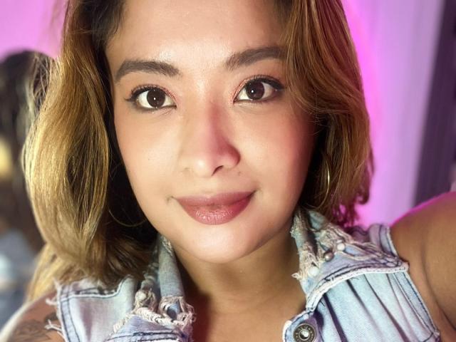 BellaLumi with brown eyes, medium skin, and wavy light brown hair, wearing a denim sleeveless top, smiling with pinkish lipstick, and small hoop earrings, looking directly at camera.