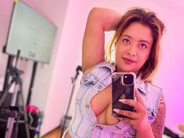 BellaLumi with her hand in her hair, wearing a cut-out denim vest that reveals her large tits, taking a selfie in a room with pink lighting.