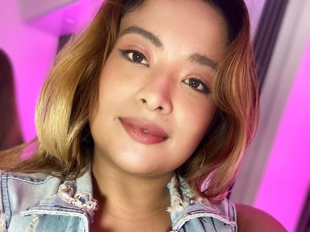 BellaLumi has light brown skin, shoulder-length blonde hair, and wears a denim jacket. She has full lips, subtle makeup, and a slight smile. Her eyes are looking at the camera.
