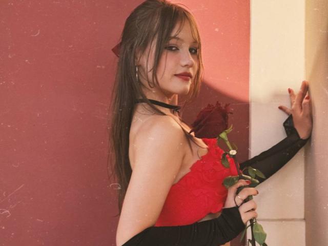 Bubochka with long brown hair, red dress, black choker, holding red rose, leaning against wall, looking over shoulder, smiling slightly, pink and white background, right arm raised.