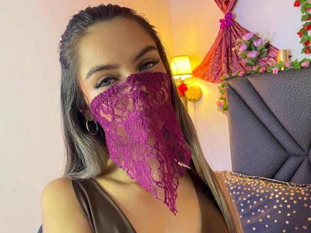 DalilaAliX with long brown hair, wearing a purple lace face mask, brown dress, and hoop earrings, looking at camera, bed with dark headboard and pink floral decor in background.