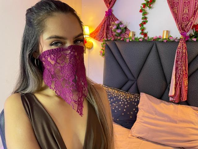 DalilaAliX with long brown hair, wearing a purple lace mask covering her mouth and nose, brown dress, and hoop earrings, sitting on a bed with pink and gold pillows.