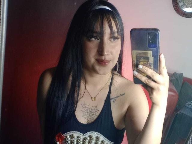 Chloey69, con cabello negro, vistiendo una camiseta sin mangas negra, sosteniendo un teléfono, mostrando un tatuaje de telaraña en el pecho y un pequeño tatuaje de corazón en el lado derecho..