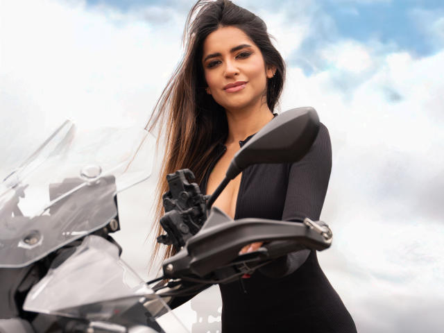 SofyaLane, avec de longs cheveux bruns, une robe noire et un léger sourire, se tient devant une moto, le bras droit sur le guidon, regardant vers le bas, un ciel clair en arrière-plan..