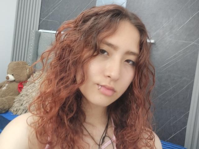 MisuGisorg has curly red hair, a nose piercing, and a slight smile, wearing a pink top, with a brown teddy bear on the left.