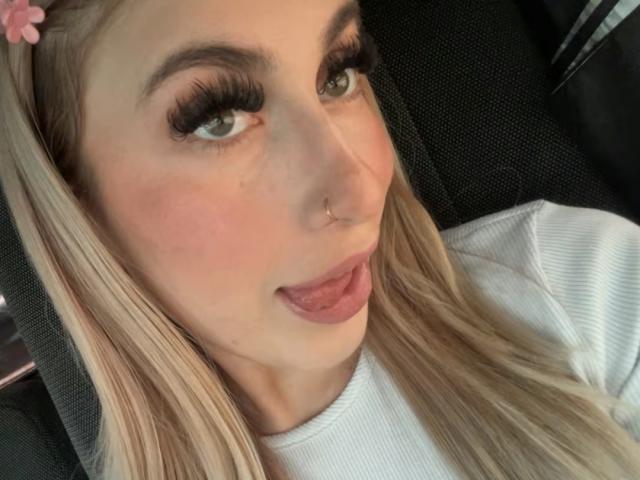 SammyViolet with long blonde hair, green eyes, and pink tongue out, wearing a white ribbed shirt, has a pink flower hairpin, and a nose ring, looking playful.
