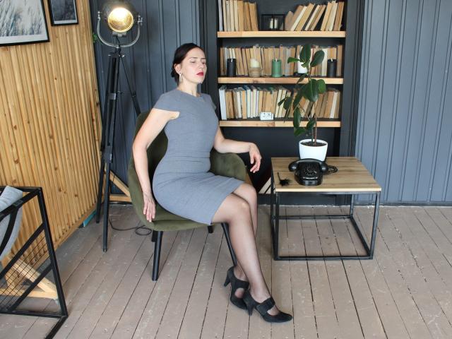 KarinaSwan with black hair, red lipstick, gray dress, black heels, and sheer pantyhose, sitting on green chair, arms relaxed, books and potted plant on wooden floor.