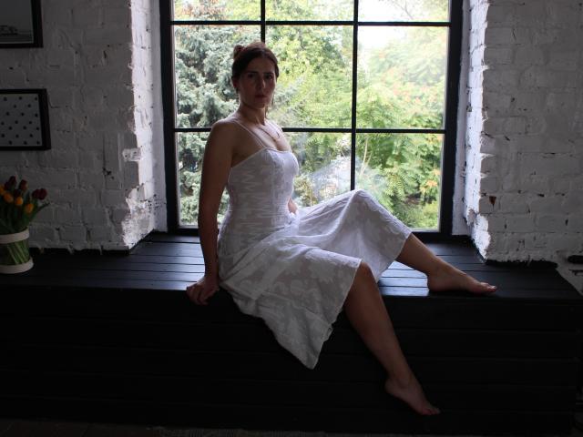 KarinaSwan, barefoot, sitting on black windowsill, white floral dress, tan skin, brown hair, small tits, looking out window, green forest outside, white brick wall, potted tulips.