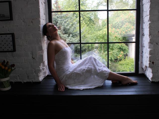KarinaSwan is sitting on a black window ledge, wearing a white, sheer dress, with her back against the ledge, head tilted up, and legs stretched out, looking sexy.