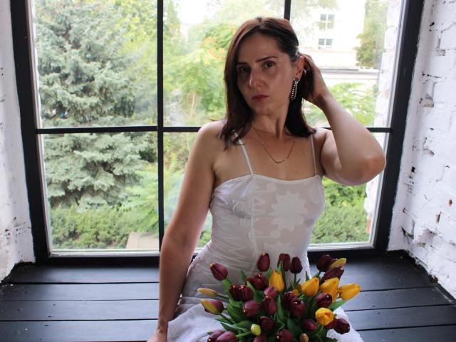 KarinaSwan with medium tits, wearing a white star-patterned dress, sitting by a window, hand in brown hair, looking serious, holding a bouquet of red and yellow tulips.