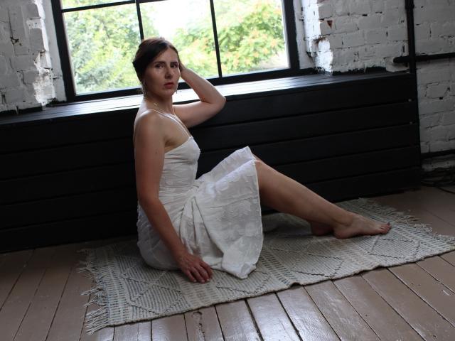 KarinaSwan with small tits, sitting on a rug, wearing a white dress, barefoot, hand on head, looking away, in a room with wooden floors and a window.