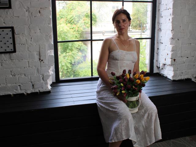 KarinaSwan, white dress, sitting on black window ledge, holding bouquet of yellow, purple, and pink tulips, white brick wall, black window frame, short brown hair, calm expression.
