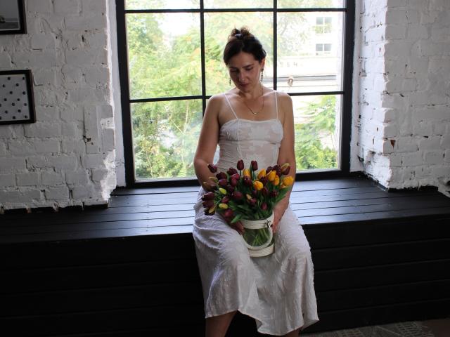 KarinaSwan with small tits, wearing a white dress, sitting on a black wooden window ledge, holding a bouquet of colorful tulips, looking down, in a white brick room.