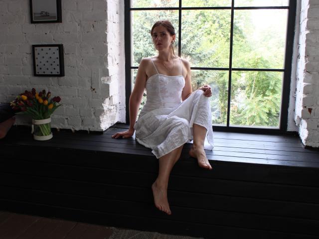 KarinaSwan sits barefoot on a black window ledge, wearing a white, strapless dress, her tits visible, with her legs crossed, looking outside, in a bright room.