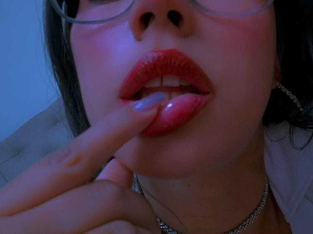CarolineWill's face with glossy red lips, finger gently touching her pink-tinted lower lip, wearing glasses, silver necklace, and a white shirt, blue lighting.