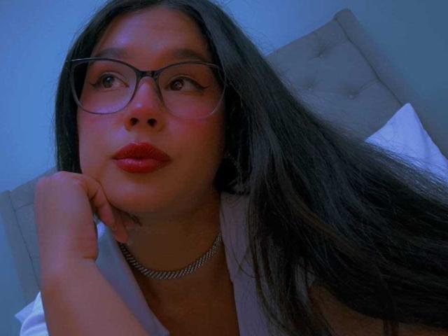 CarolineWill with long black hair, glasses, red lipstick, white shirt, and silver chain, resting her chin on her hand, looking to the side, in a blue-lit room.