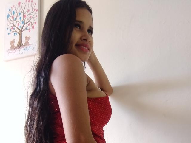 ScarlettRoss69, with long black hair, smiling, wearing a red polka dot strapless top, showing her tits, hand on her head, standing against a white wall, looking over her shoulder.