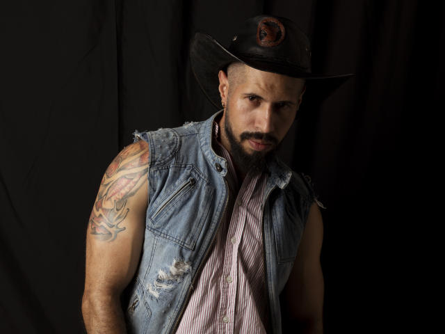 Tattooed Yen69 with a beefy arm, wearing a dirty denim vest, striped shirt, and black cowboy hat, staring with a serious face. Dark background.