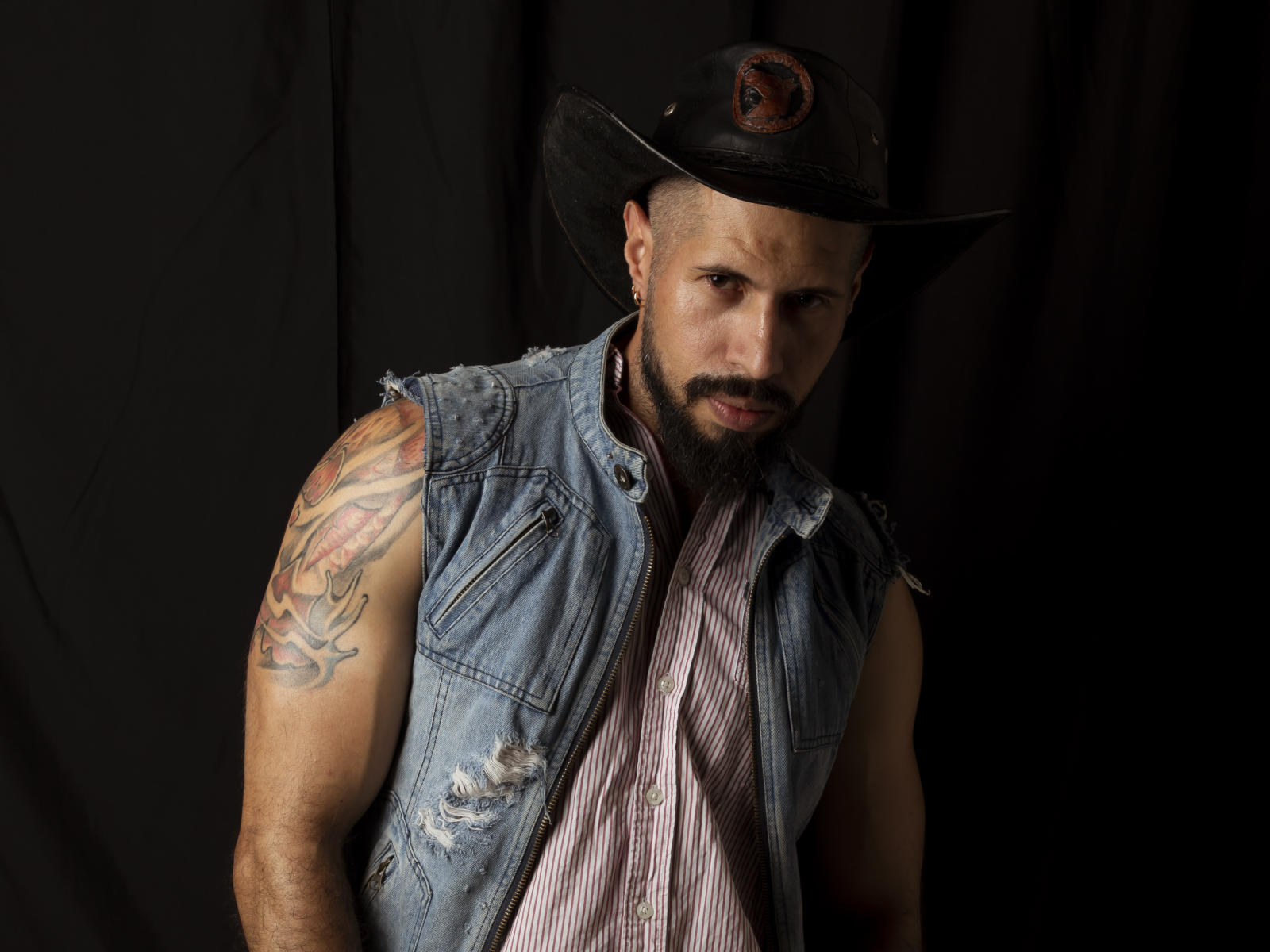 Tattooed Yen69 with a beefy arm, wearing a dirty denim vest, striped shirt, and black cowboy hat, staring with a serious face. Dark background.