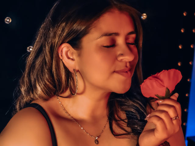 MelindaBoobs with closed eyes, holding a pink rose to her nose. She has wavy brown hair, a nose ring, hoop earrings, and a necklace. She's wearing a black top.