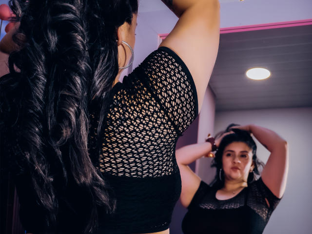MelindaBoobs with long, curly black hair, wearing a black mesh top, standing in front of a mirror, adjusting her hair with both hands, reflecting in the mirror.