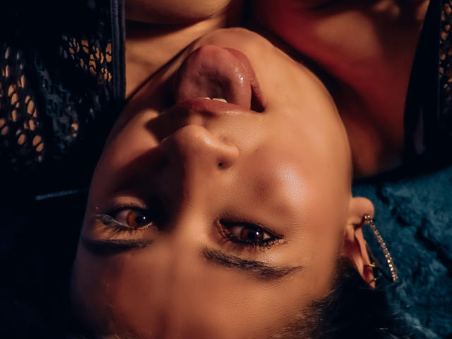 MelindaBoobs' face is upside down with her tongue out, brown eyes looking up, wearing black lace, and a silver earring in her left ear, on a blue textured surface.