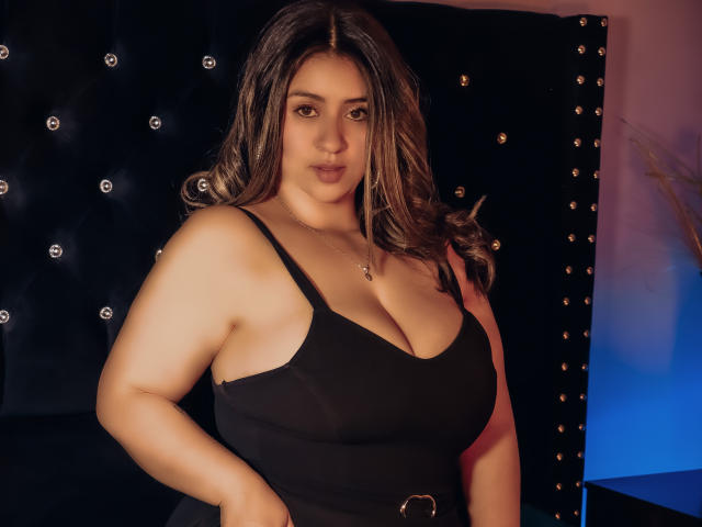 MelindaBoobs with large tits, wearing a black tank top, has wavy brown hair, a gold necklace, and a confident expression, standing against a tufted black wall.
