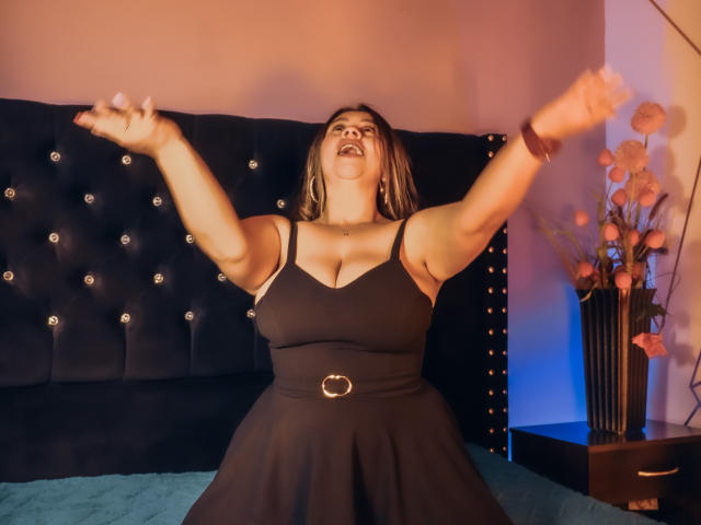 MelindaBoobs with arms raised, mouth open, wearing a black dress with a belt, showing her tits, in a bedroom with a tufted black headboard and blue bed.