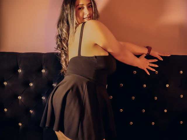 MelindaBoobs with long, wavy hair, wearing a black dress, leaning on a black tufted couch, showing her ass, smiling at the camera, in a warmly lit room.