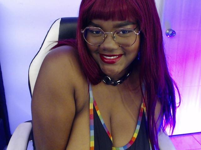 RoseBeckers with red hair, glasses, and red lipstick, smiling, showing large tits in a striped tank top, sitting in a white and black chair. Choker necklace. Purple lighting.