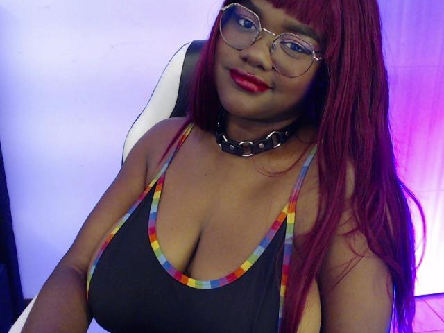 RoseBeckers with red hair, glasses, and red lipstick, wearing a black tank top with rainbow trim, and a black choker, sitting in a chair, showing her big boobs.