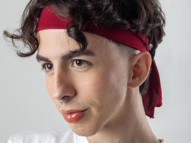 BrahmBelial, con cabello castaño rizado, una diadema roja, un piercing en forma de estrella en la nariz y labios anaranjados brillantes, mirando ligeramente hacia un lado. Piel clara, ojos marrones, camisa blanca..