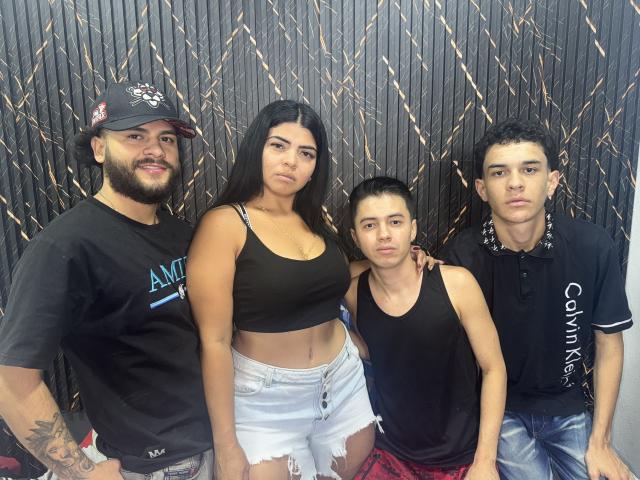 Four people, MeganeGtGangs with dark hair, black crop top, and white shorts, standing with a bearded man, a girl, and a boy in black shirts, against a dark wall.