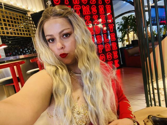 SamanthaGold with long blonde wavy hair, red lipstick, and a gold-embellished top, taking a selfie in a brightly lit restaurant, showing off her tits and a cute face.