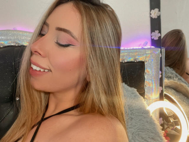JennyLawry with blonde hair, pink eyeshadow, black eyeliner, smiling, showing her ass, wearing a black halter top, reflection in mirror, ring light, soft pink and purple lights.