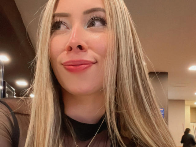 JennyLawry's face, blonde hair, light skin, subtle makeup, pink lips, slight smile, black sheer top, visible eyelashes, looking up, indoor setting, casual selfie, soft lighting.