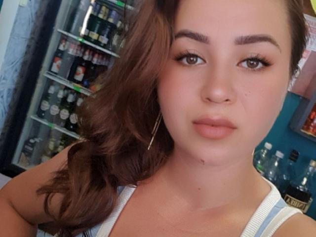 HottieBLove with brown wavy hair, wearing a white tank top, has full lips, subtle makeup, and brown eyes, looking directly at the camera with a slight pout.