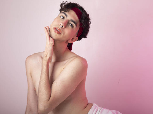 BrahmBelial, sem camisa, com cabelo castanho encaracolado, uma faixa vermelha na cabeça e pele clara, toca o pescoço, mostrando um peito magro e peludo, e um fundo rosado. Os lábios de BrahmBelial estão ligeiramente entreabertos..