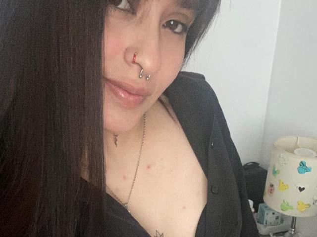 MeghaanRoux with long black hair, nose ring, and lip ring, wearing a black shirt, showing a tattoo near her tits, and a necklace, in a selfie.