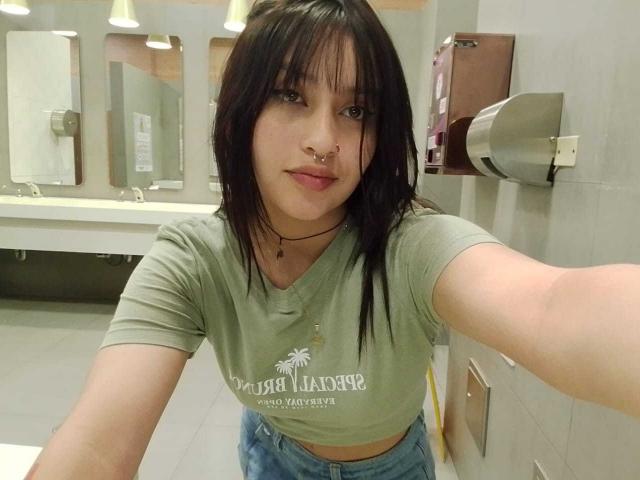 MeghaanRoux with black hair, green t-shirt, nose ring, and jeans, taking a selfie in a bathroom, showing a slight midriff, and a confident expression.