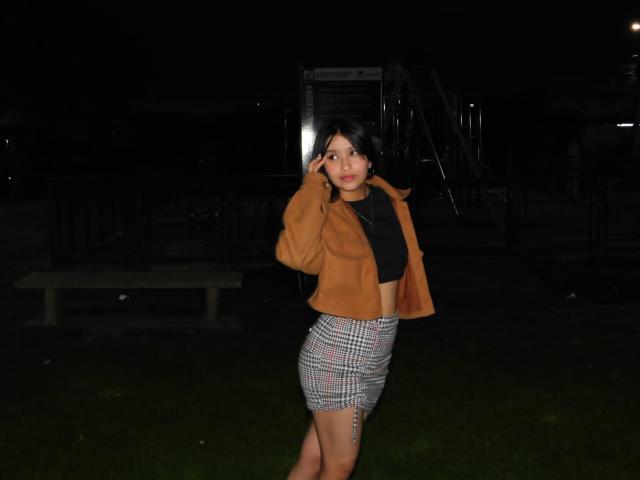 MeghaanRoux stands in a dark night scene, wearing a brown suede jacket, black crop top, and a checkered skirt, with one hand touching her head and a small tattoo on her thigh.