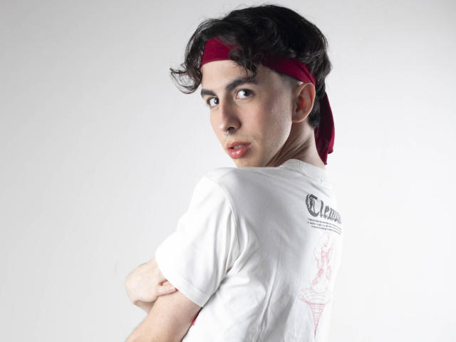 BrahmBelial con cabello rizado oscuro, diadema roja, camisa blanca, brazos cruzados, mirando por encima del hombro, boca ligeramente abierta, piel clara, expresión suave, fondo gris..