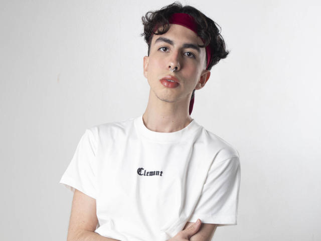 BrahmBelial with curly brown hair, red headband, white t-shirt, and arms crossed, looking serious, has slight facial stubble, and is wearing red lipstick.