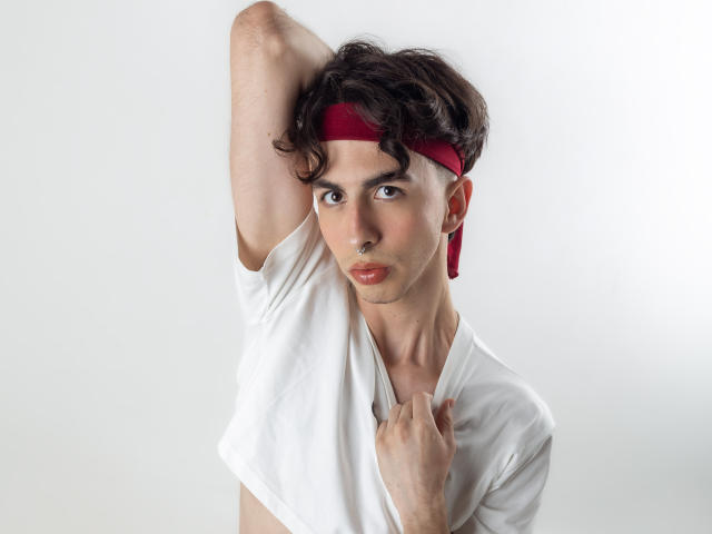 BrahmBelial met een rode hoofdband, een wit shirt en een serieuze uitdrukking, die zijn arm omhoog steekt en zijn shirt vasthoudt met een hand, waarbij een slank, aantrekkelijk gezicht en borst zichtbaar zijn..