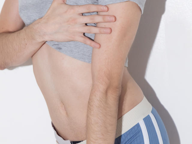 Het slanke, bleke torso van BrahmBelial, met zichtbare armhaar, draagt een grijze shirt en blauwe korte broek met witte strepen, en heeft een hand op hun zij. Het watermerk van de foto is zichtbaar..